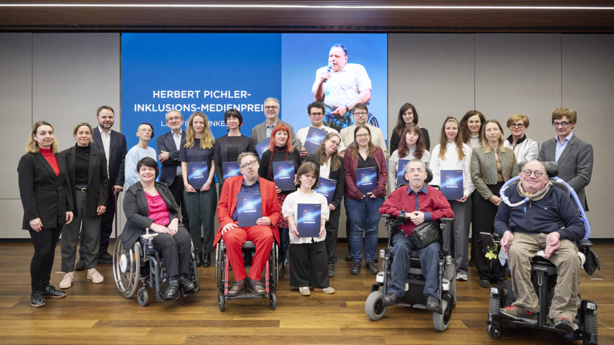 Gruppenfoto aller Gewinner*innen des Herbert-Pichler-Inklusions-Medienpreises 2026 auf einer Bühne am ORF Mediencampus. Mehrere Personen stehen und sitzen nebeneinander, einige nutzen Rollstühle, viele halten ihre Auszeichnungen. Im Hintergrund ist eine große Leinwand mit dem Veranstaltungstitel und einem Bild von Herbert Pichler zu sehen.