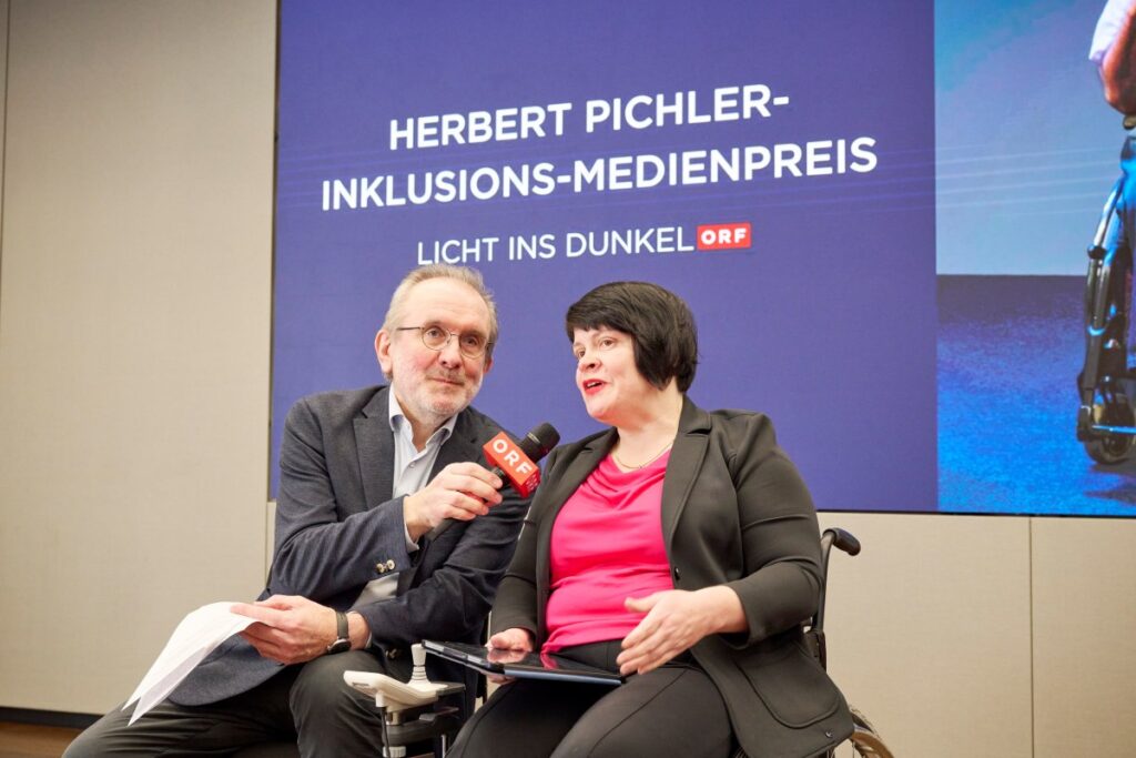 Eine Frau und ein Mann sitzen auf einer Bühne. Die Frau nutzt einen Rollstuhl, der Mann hält ein ORF-Mikrofon in der Hand. Im Hintergrund "Herbert Pichler-Inklusions-Medienpreis Licht ins Dunkel" und ein Foto von Herbert Pichler
