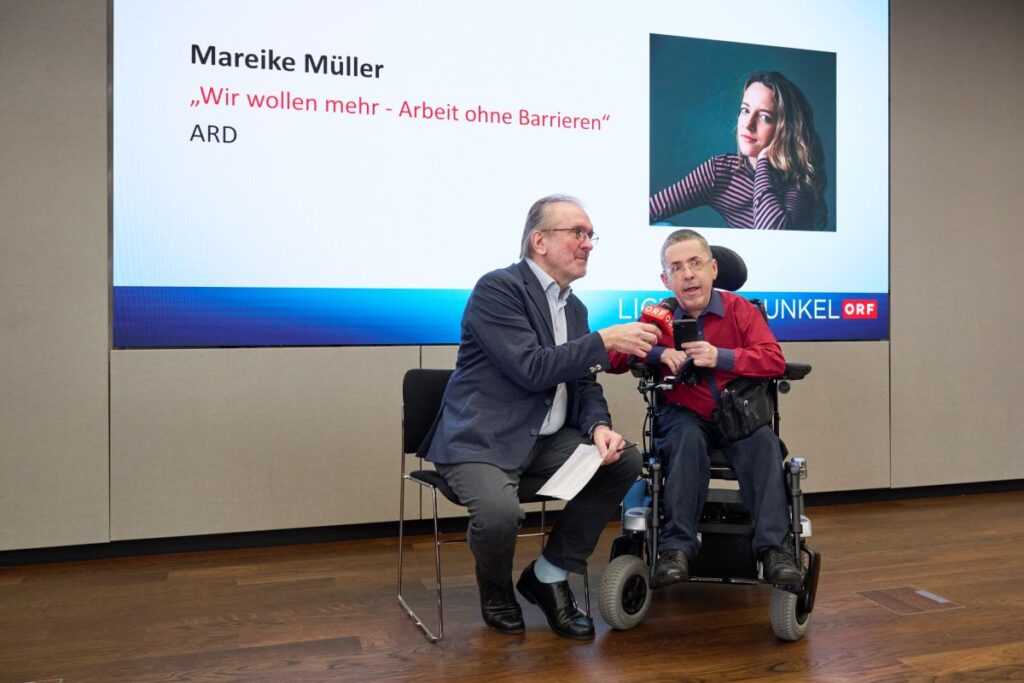 2 Männer sitzen auf einer Bühne. Einer davon nutzt einen Rollstuhgl, der andere hält ein ORF-Mikrofon in der Hand. Im Hintergrund "Herbert Pichler-Inklusions-Medienpreis Licht ins Dunkel" und ein Foto von Herbert Pichler