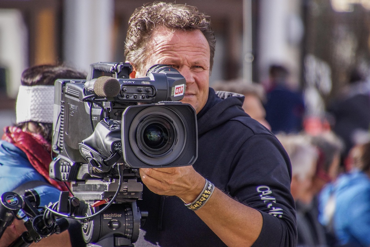 Ein Kameramann filmt mit einer professionellen Fernsehkamera mit ORF-Logo bei einer Veranstaltung im Freien. Im Hintergrund sind Menschen unscharf zu sehen. Foto: Pixabay/rottonara