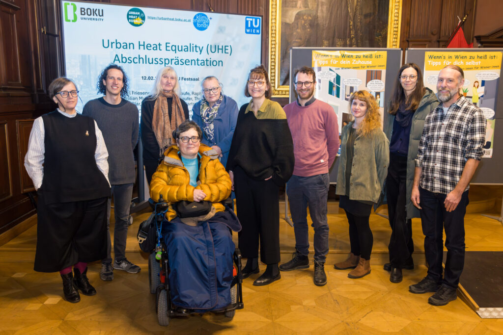 Gruppe von Menschen aus Projektteam und beteiligten Organisationen des Forschungsprojekts Urban Heat Equality steht in einem Raum vor einer Präsentationswand mit dem Titel „Urban Heat Equality Abschlusspräsentation“. Eine Person sitzt im Rollstuhl im Vordergrund, die anderen stehen daneben. Im Hintergrund sind Logos von beteiligten Institutionen sowie Informationsplakate zu Hitzeschutzmaßnahmen in Wien zu sehen.