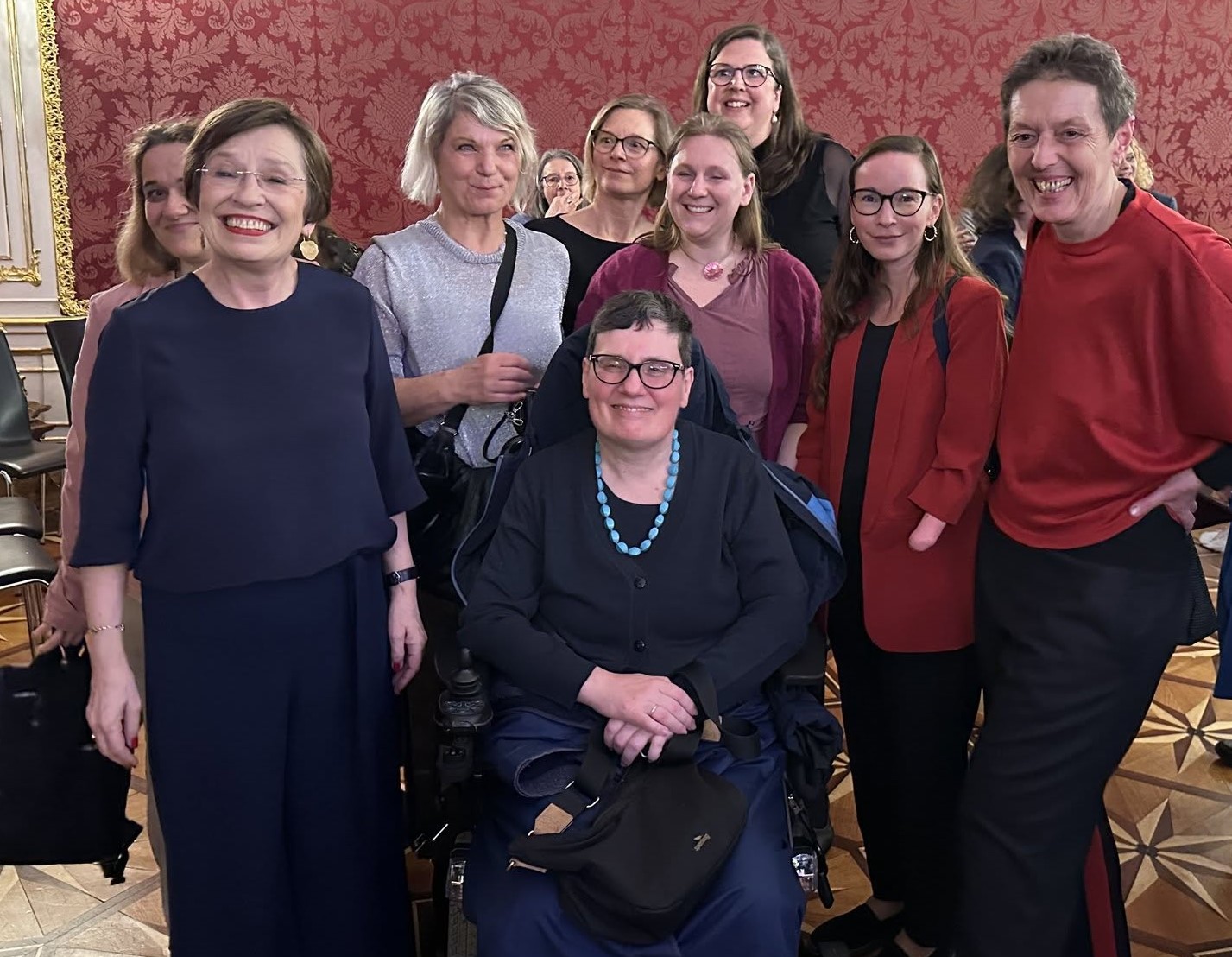 Gruppenfoto in einem prunkvollen Innenraum mit roter, ornamentierter Wandtapete und Parkettboden. Mehrere Frauen stehen dicht beieinander und lächeln in die Kamera. Im Vordergrund sitzt eine Frau mit kurzen dunklen Haaren und Brille in einem Rollstuhl. Sie trägt ein dunkles Outfit und eine auffällige türkisfarbene Halskette. Neben ihr stehen mehrere Frauen unterschiedlichen Alters, einige mit Brille, in überwiegend dunkler Kleidung mit einzelnen roten Akzenten. Die Gruppe wirkt entspannt und freundlich, einige lehnen sich leicht zueinander. Im Hintergrund sind weitere Personen teilweise zu sehen.