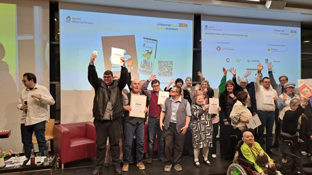 Mehrere ausgezeichnete Personen stehen gemeinsam auf einer Bühne und halten ihre Urkunden in die Höhe. Einige heben die Hände und freuen sich über die Auszeichnung. Im Vordergrund sitzt unter anderem eine Person im Rollstuhl. Im Hintergrund sind Präsentationsfolien zum Literaturpreis Ohrenschmaus 2026 zu sehen.