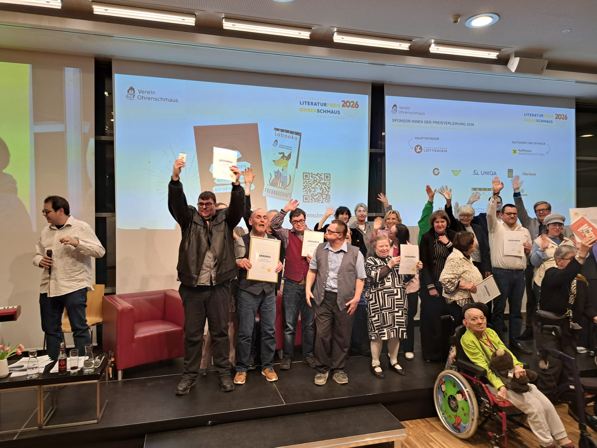 Mehrere ausgezeichnete Personen stehen gemeinsam auf einer Bühne und halten ihre Urkunden in die Höhe. Einige heben die Hände und freuen sich über die Auszeichnung. Im Vordergrund sitzt unter anderem eine Person im Rollstuhl. Im Hintergrund sind Präsentationsfolien zum Literaturpreis Ohrenschmaus 2026 zu sehen.