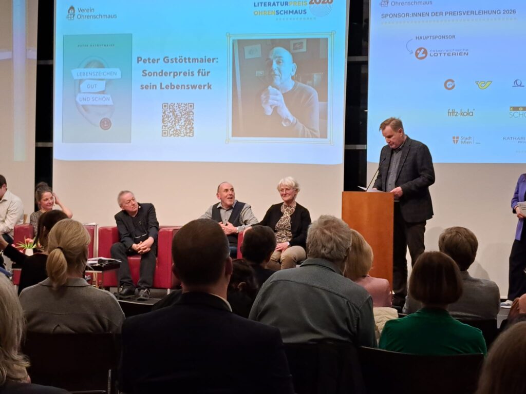 Ein Mann steht an einem Rednerpult und hält eine Laudatio, während mehrere Personen auf einer Bühne sitzen. Im Hintergrund ist eine Präsentation zum Sonderpreis für das Lebenswerk von Peter Gstöttmaier zu sehen. Das Publikum sitzt im Vordergrund und verfolgt die Veranstaltung.
