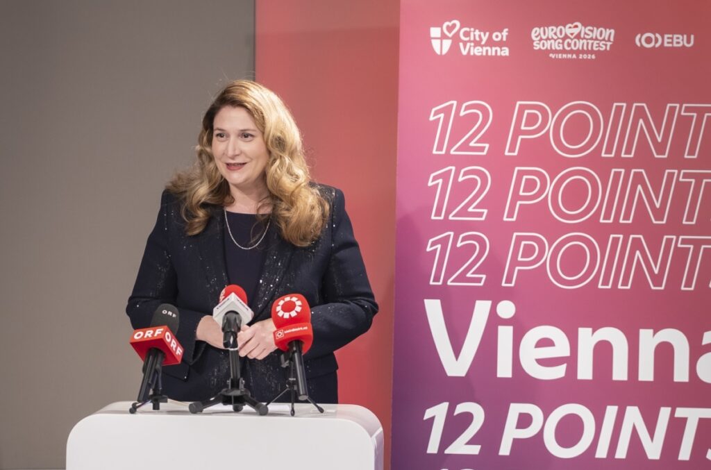 Eine Frau steht bei einer Pressekonferenz hinter einem Rednerpult und spricht in mehrere Mikrofone. Hinter ihr ist ein großes rosa Banner mit den Logos „City of Vienna“, „Eurovision Song Contest Vienna 2026“ und dem Schriftzug „12 Points Vienna“ zu sehen.