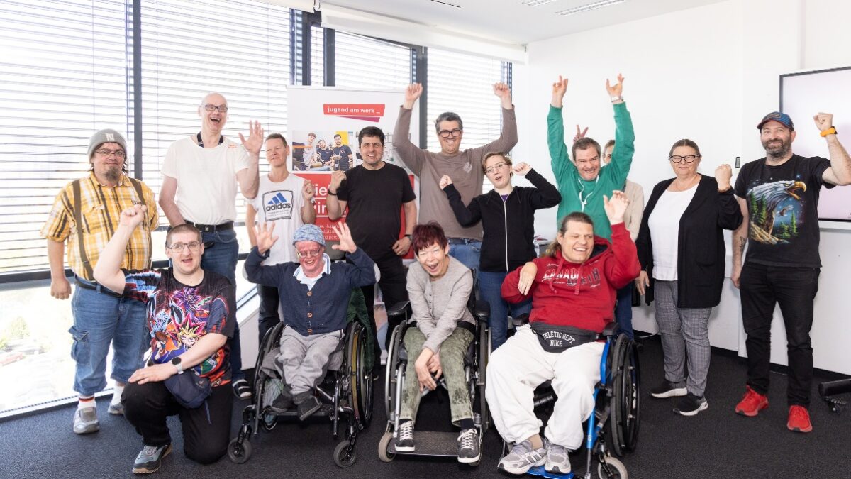 Gruppe von Menschen mit und ohne Behinderungen in einem hellen Raum. Einige Personen sitzen im Rollstuhl, andere stehen dahinter. Viele heben die Arme und lächeln. Im Hintergrund steht ein Roll-up mit dem Logo „jugend am werk“.