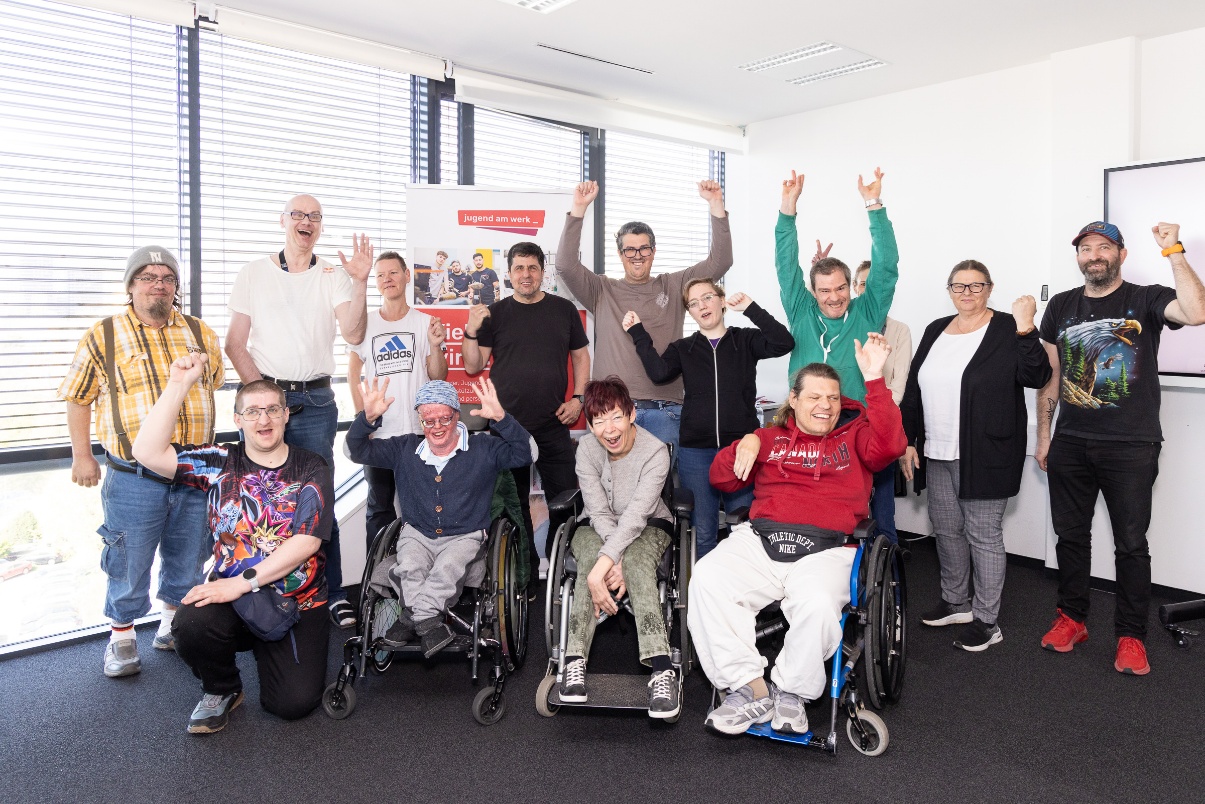Gruppe von Menschen mit und ohne Behinderungen in einem hellen Raum. Einige Personen sitzen im Rollstuhl, andere stehen dahinter. Viele heben die Arme und lächeln. Im Hintergrund steht ein Roll-up mit dem Logo „jugend am werk“.