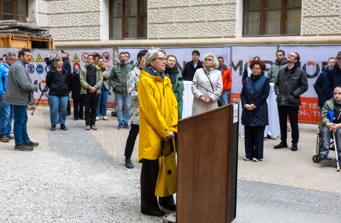 Eine Frau spricht an einem Rednerpult im Innenhof des Naturhistorischen Museums Wien bei der Eröffnung des neuen barrierefreien Besucherlifts. Mehrere Personen stehen im Hintergrund und hören zu.
