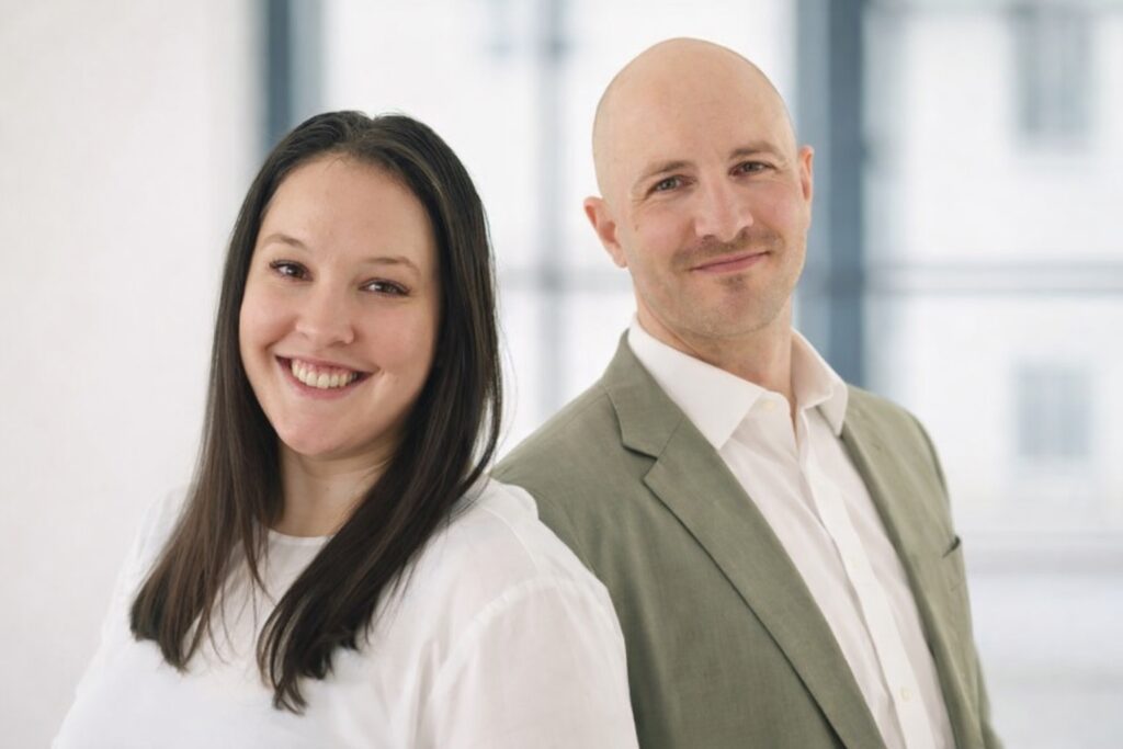 Porträtfoto der Referent*innen Mag.a Victoria Biber und Felix Steigmann. Victoria Biber trägt ein weißes Oberteil und lächelt in die Kamera. Neben ihr steht Felix Steigmann mit hellem Sakko und weißem Hemd. Beide sind vor einem hellen, leicht verschwommenen Hintergrund fotografiert.