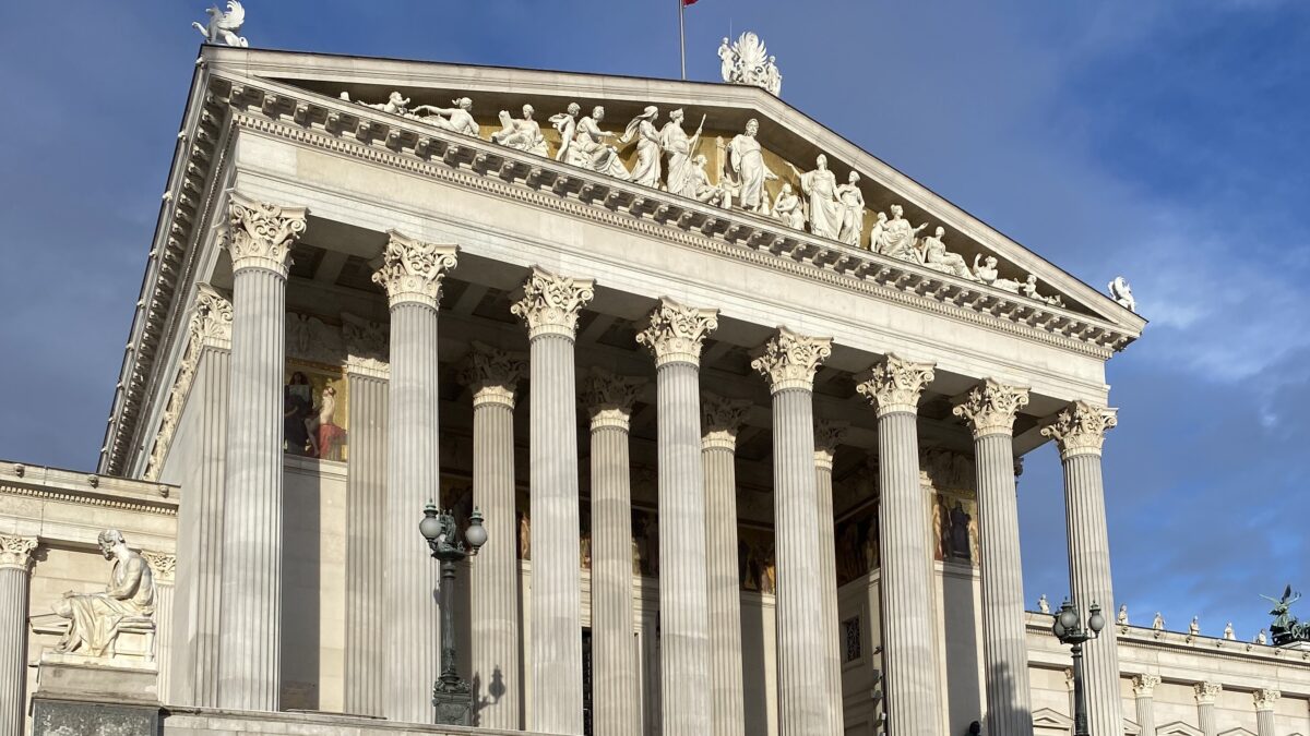 Frontansicht des österreichischen Parlamentsgebäudes in Wien mit hohen Säulen, Figuren am Giebel und einer roten Flagge auf dem Dach vor blauem Himmel.