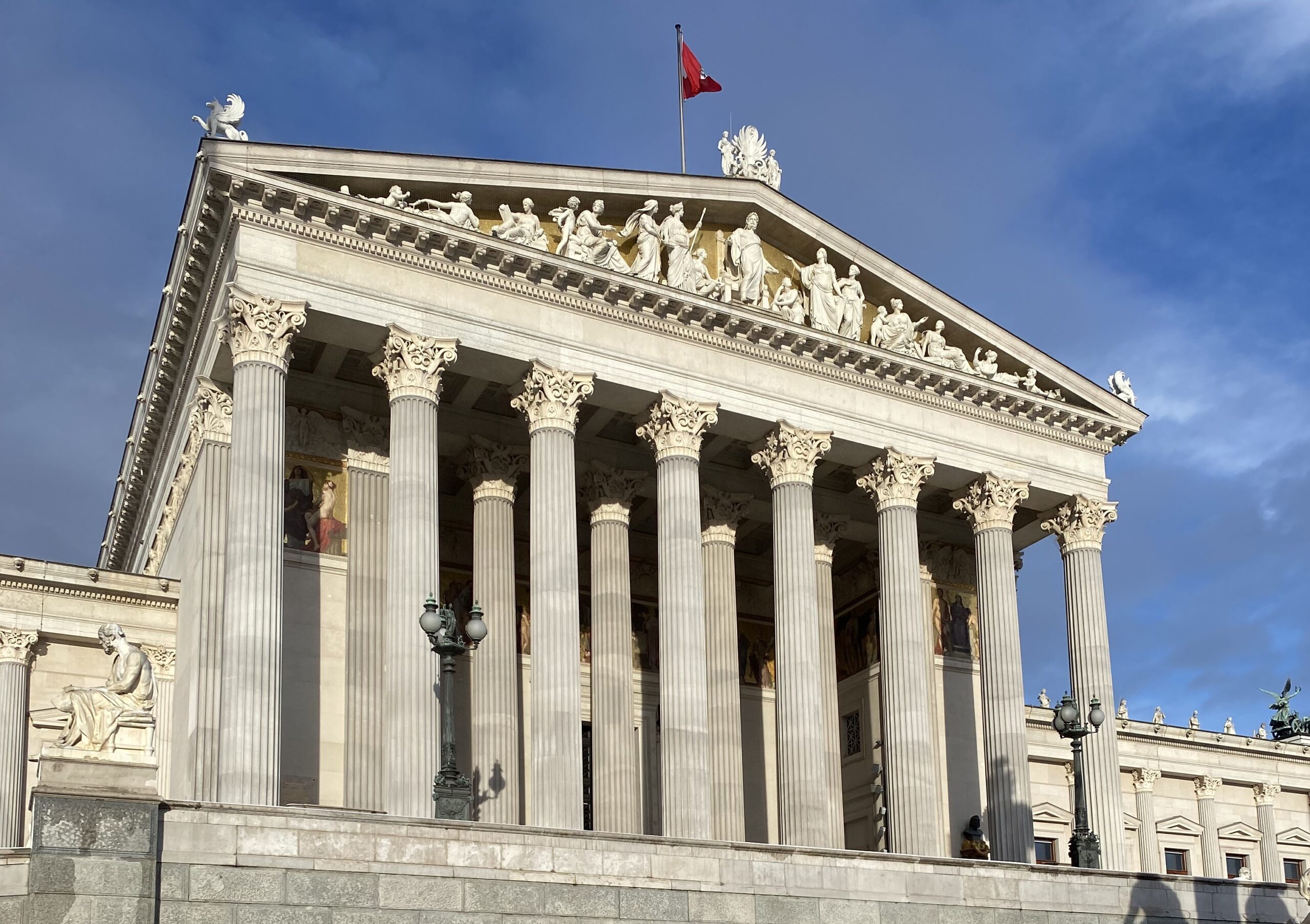 Frontansicht des österreichischen Parlamentsgebäudes in Wien mit hohen Säulen, Figuren am Giebel und einer roten Flagge auf dem Dach vor blauem Himmel.