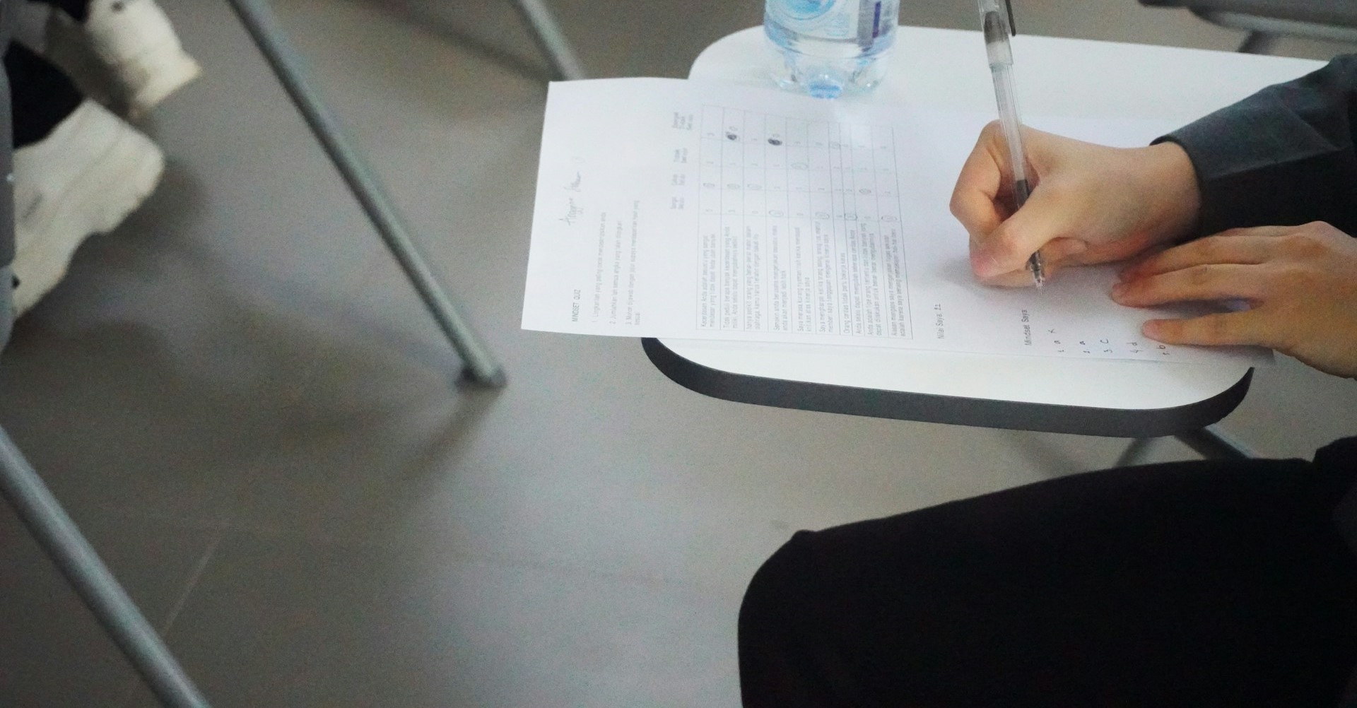 Person sitzt in einem Klassenraum und füllt einen Testbogen auf einem kleinen Schultisch aus; daneben steht eine Wasserflasche.