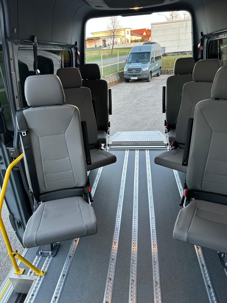 Innenraum eines rollstuhlgerechten Kleinbusses mit seitlich angeordneten Sitzen, Schienensystem zur Rollstuhlsicherung und ausgeklappter Rampe am Heck, Blick nach außen.