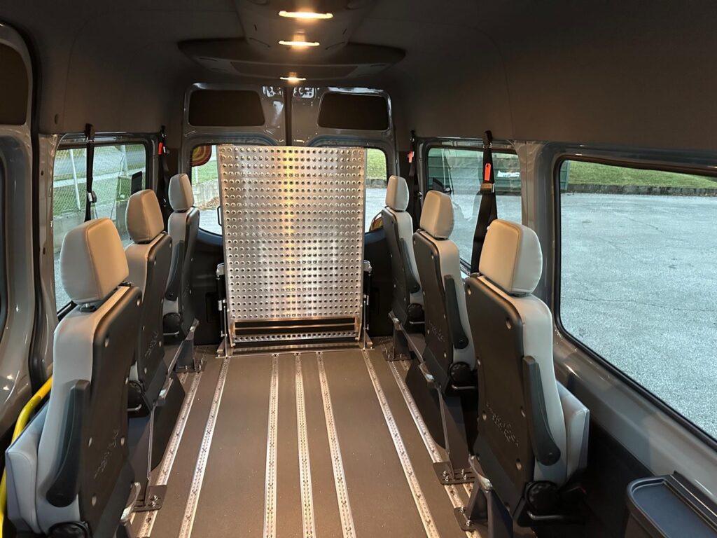 Innenraum eines rollstuhlgerechten Kleinbusses mit Klappsitzen, Schienensystem zur Rollstuhlsicherung und aufgestellter Rampe im hinteren Bereich.