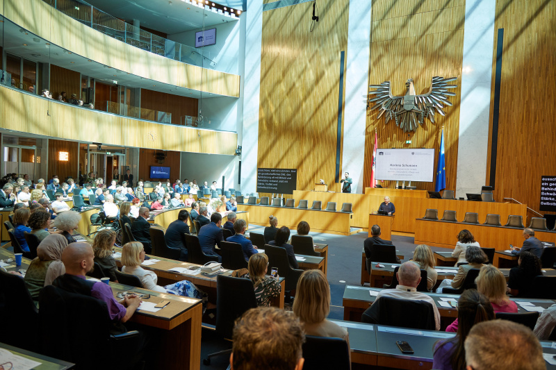 Blick in den Sitzungssaal des österreichischen Parlaments während einer Veranstaltung. Sozialministerin Korinna Schumann spricht am Rednerpult vor zahlreichen Besucher*innen.