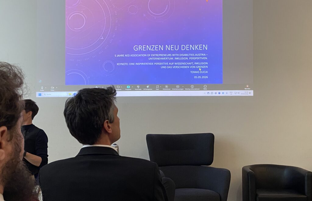 Publikum bei der Jubiläumsveranstaltung „5 Jahre AED“ während einer Präsentation mit dem Titel „Grenzen neu denken“. Auf eine große Leinwand wird die Keynote von Tomas Ducai projiziert. Im Vordergrund sitzen Gäste im Veranstaltungsraum und verfolgen den Vortrag.