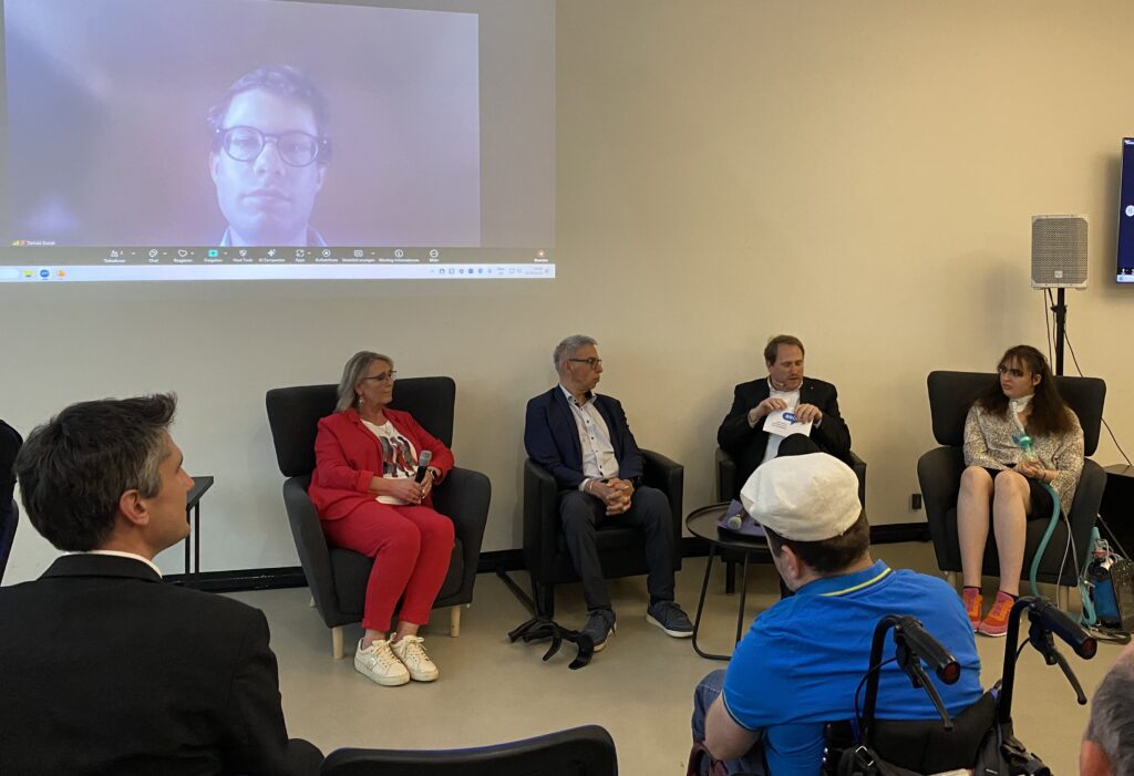 Podiumsdiskussion bei der Jubiläumsveranstaltung „5 Jahre AED“ mit mehreren Teilnehmern auf einer Bühne vor Publikum. Eine Moderatorin steht mit Mikrofon neben den sitzenden Diskussionsteilnehmern. Im Hintergrund sind Präsentationsflächen mit dem AED-Logo sowie Veranstaltungstechnik zu sehen. Im Publikum sitzt unter anderem ein Rollstuhlnutzer.
