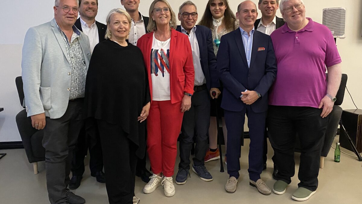 Gruppenfoto bei der Jubiläumsveranstaltung „5 Jahre AED“ mit mehreren Personen aus Vorstand, Beirat und Umfeld der Association of Entrepreneurs with Disabilities Austria in einem Veranstaltungsraum. Die Teilnehmenden stehen nebeneinander und lächeln in die Kamera. In der Mitte steht eine Frau in rotem Hosenanzug, rundherum mehrere Männer und eine weitere Frau in dunkler Kleidung. Im Hintergrund sind Sitzmöbel und Veranstaltungstechnik zu sehen.