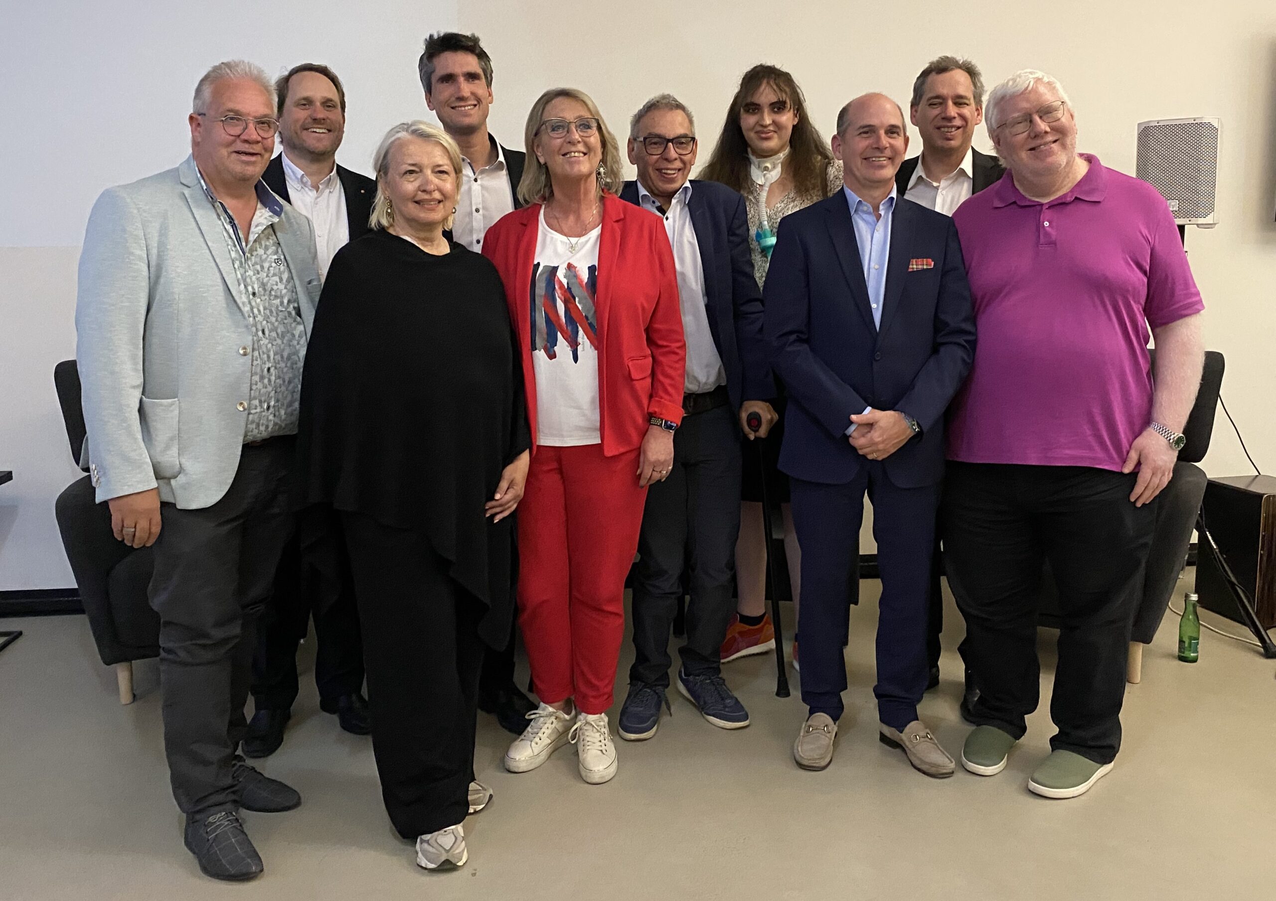 Gruppenfoto bei der Jubiläumsveranstaltung „5 Jahre AED“ mit mehreren Personen aus Vorstand, Beirat und Umfeld der Association of Entrepreneurs with Disabilities Austria in einem Veranstaltungsraum. Die Teilnehmenden stehen nebeneinander und lächeln in die Kamera. In der Mitte steht eine Frau in rotem Hosenanzug, rundherum mehrere Männer und eine weitere Frau in dunkler Kleidung. Im Hintergrund sind Sitzmöbel und Veranstaltungstechnik zu sehen.