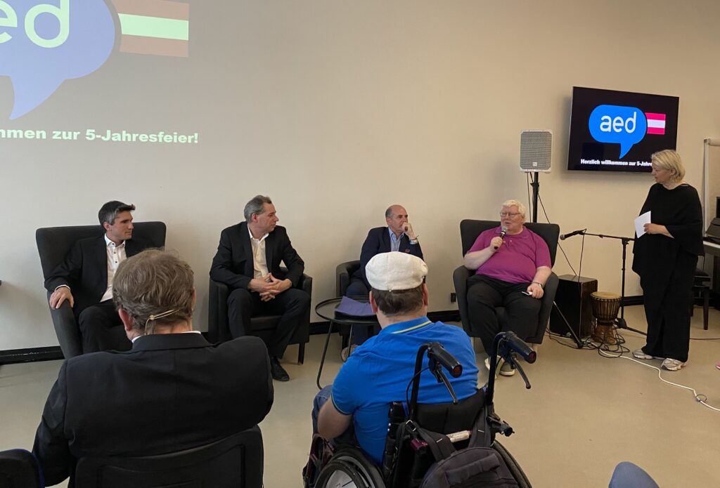 Podiumsdiskussion bei der Jubiläumsveranstaltung „5 Jahre AED“ mit mehreren Teilnehmern auf einer Bühne vor Publikum. Eine Moderatorin steht mit Mikrofon neben den sitzenden Diskussionsteilnehmern. Im Hintergrund sind Präsentationsflächen mit dem AED-Logo sowie Veranstaltungstechnik zu sehen. Im Publikum sitzt unter anderem ein Rollstuhlnutzer.