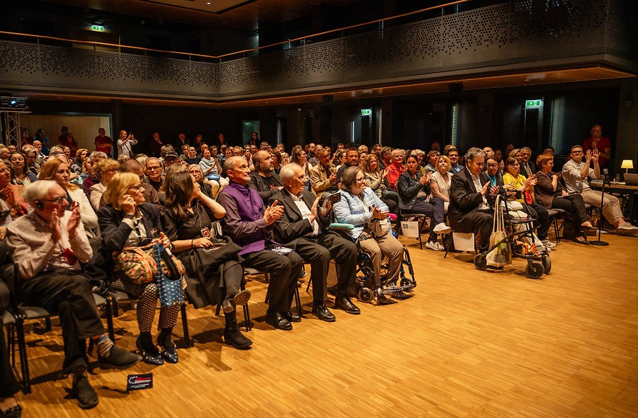 Publikum im Veranstaltungssaal des Filmfestivals ART WITHOUT LIMIT. Zahlreiche Besucher*innen sitzen auf Stühlen, einige Personen nutzen Rollstühle oder Rollatoren. Die Menschen applaudieren aufmerksam in einem modern beleuchteten Saal mit Balkon und Holzfußboden.