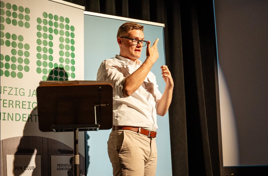 David Obermaier gebärdet auf der Bühne des Filmfestivals ART WITHOUT LIMIT. Er trägt ein weißes Hemd und steht vor einem Banner des Österreichischen Behindertenrats. Seine Hände sind während der Gebärdensprache erhoben.