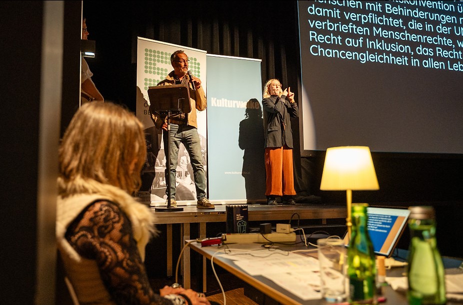 Klaus Widl steht bei einer Veranstaltung des Filmfestivals ART WITHOUT LIMIT an einem Rednerpult und spricht in ein Mikrofon. Neben ihm dolmetscht eine Gebärdensprachdolmetscherin. Im Hintergrund sind Banner sowie eine große Leinwand mit eingeblendeten Untertiteln zu sehen.