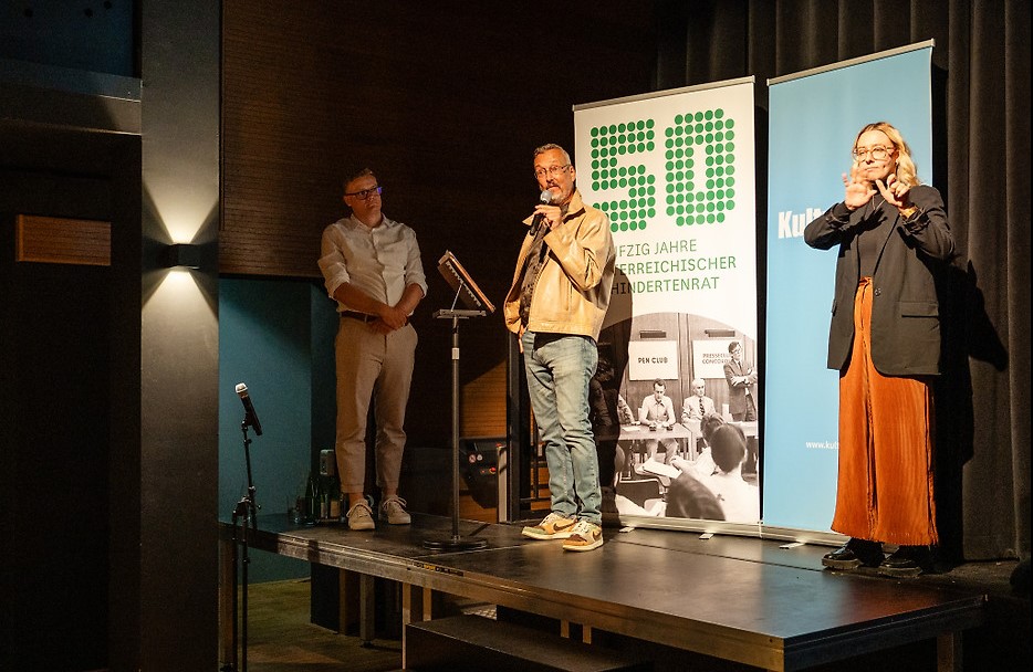 Klaus Widl steht bei einer Veranstaltung des Filmfestivals ART WITHOUT LIMIT an einem Rednerpult und spricht in ein Mikrofon. Neben ihm dolmetscht eine Gebärdensprachdolmetscherin. Im Hintergrund sind Banner sowie eine große Leinwand mit eingeblendeten Untertiteln zu sehen.