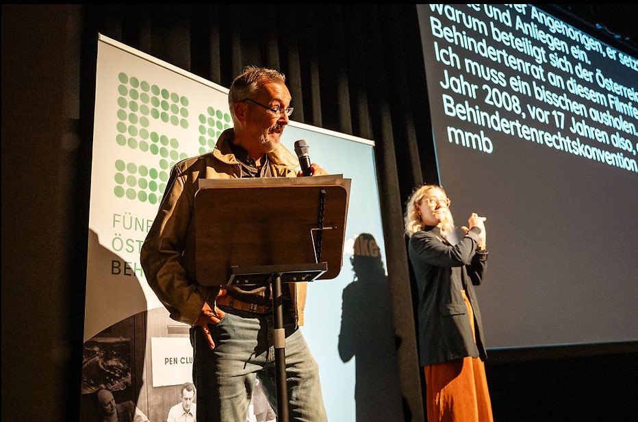Klaus Widl steht bei einer Veranstaltung des Filmfestivals ART WITHOUT LIMIT an einem Rednerpult und spricht in ein Mikrofon. Neben ihm dolmetscht eine Gebärdensprachdolmetscherin. Im Hintergrund sind Banner sowie eine große Leinwand mit eingeblendeten Untertiteln zu sehen.