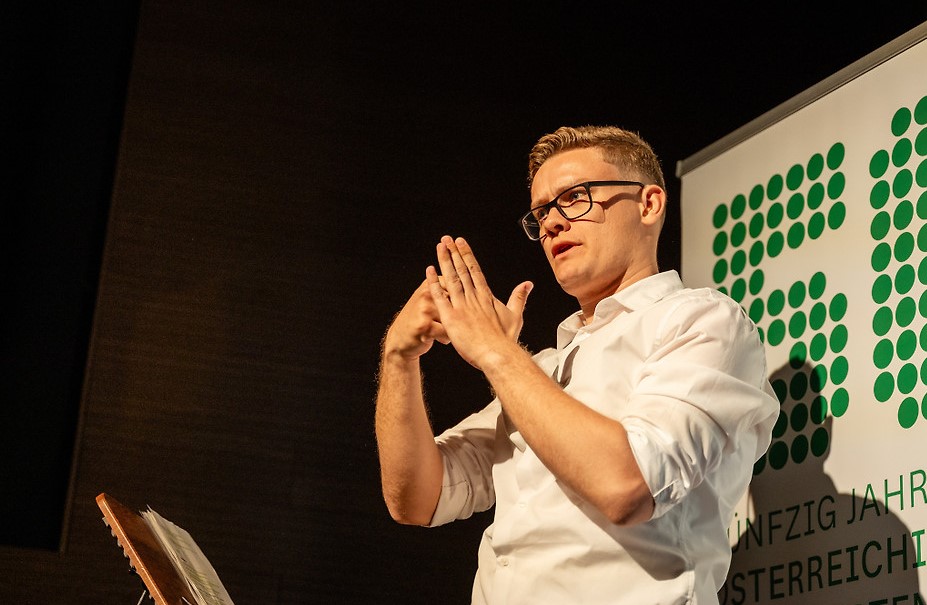 David Obermaier gebärdet auf der Bühne des Filmfestivals ART WITHOUT LIMIT. Er trägt ein weißes Hemd und steht vor einem Banner des Österreichischen Behindertenrats. Seine Hände sind während der Gebärdensprache erhoben.