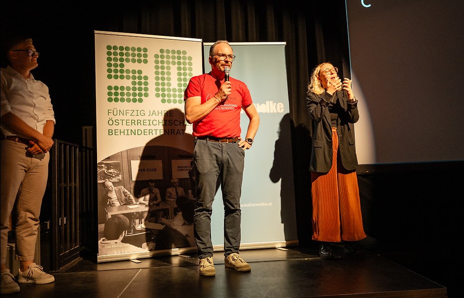 Hans-Peter Arzberger steht auf der Bühne des Filmfestivals ART WITHOUT LIMIT und spricht in ein Mikrofon. Neben ihm dolmetscht eine ÖGS-Dolmetscherin in Gebärdensprache. Im Hintergrund sind Banner des Österreichischen Behindertenrats und einer Kulturorganisation zu sehen.