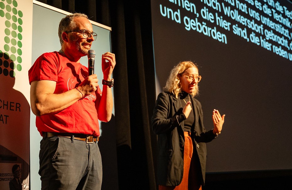 Hans-Peter Arzberger steht auf der Bühne des Filmfestivals ART WITHOUT LIMIT und spricht in ein Mikrofon. Neben ihm dolmetscht eine ÖGS-Dolmetscherin in Gebärdensprache. Im Hintergrund sind Banner des Österreichischen Behindertenrats und einer Kulturorganisation zu sehen.