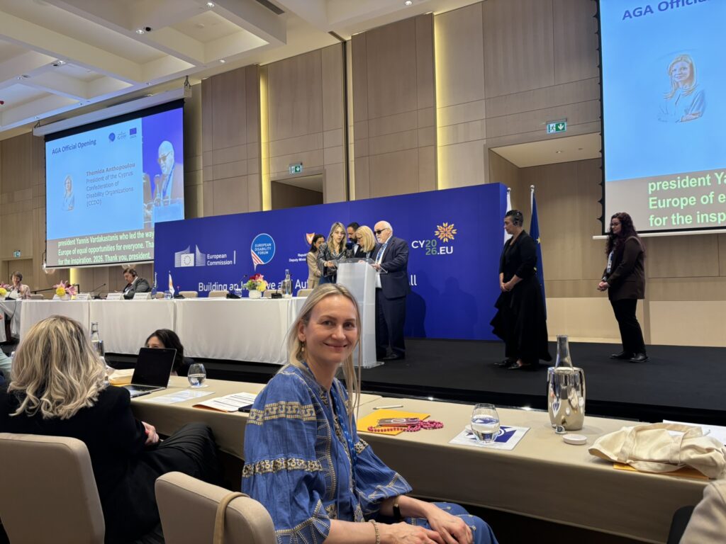 Helene Jarmer sitzt in einem Konferenzsaal bei der Generalversammlung des European Disability Forum in Zypern und blickt in die Kamera. Im Hintergrund stehen mehrere Personen auf einer Bühne vor einer großen blauen Veranstaltungswand mit Logos der Europäischen Kommission, des European Disability Forum und der zyprischen EU-Ratspräsidentschaft. Auf zwei großen Leinwänden sind Programminhalte und Redner*innen zu sehen.