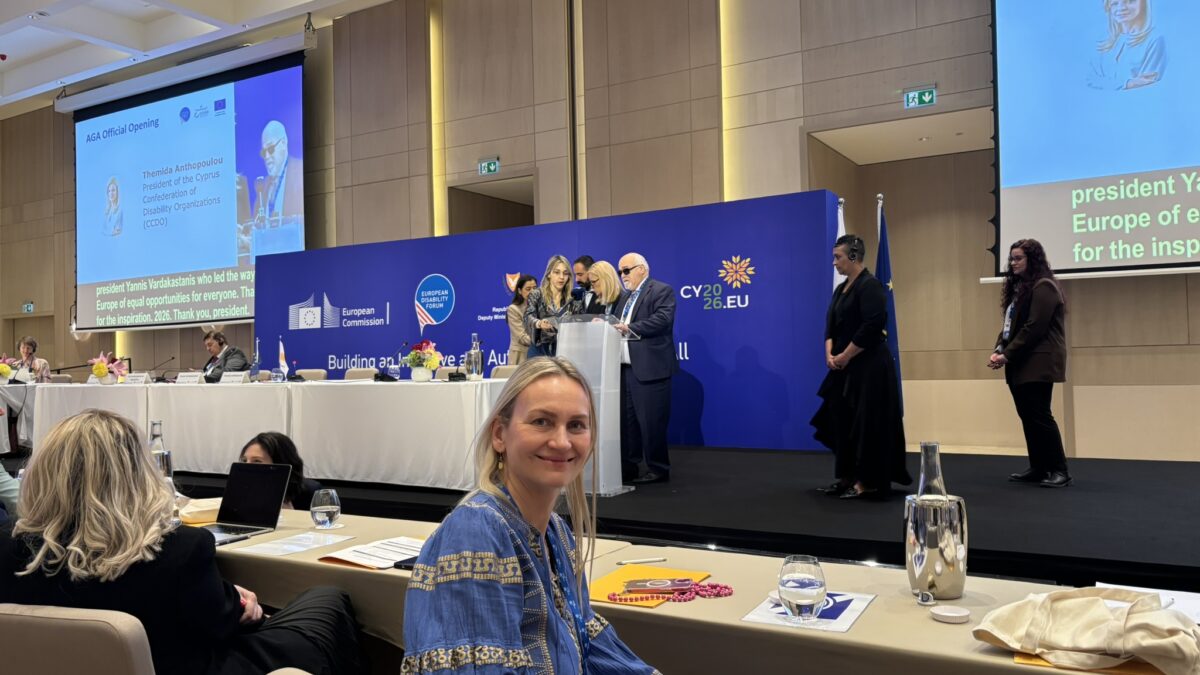 Helene Jarmer sitzt in einem Konferenzsaal bei der Generalversammlung des European Disability Forum in Zypern und blickt in die Kamera. Im Hintergrund stehen mehrere Personen auf einer Bühne vor einer großen blauen Veranstaltungswand mit Logos der Europäischen Kommission, des European Disability Forum und der zyprischen EU-Ratspräsidentschaft. Auf zwei großen Leinwänden sind Programminhalte und Redner*innen zu sehen.