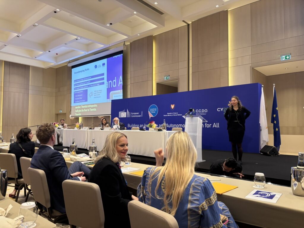 Teilnehmer*innen sitzen bei der Generalversammlung des European Disability Forum in einem Konferenzsaal in Zypern. Im Vordergrund unterhalten sich mehrere Personen an einem Tisch. Auf der Bühne spricht eine Frau vor einer großen blauen Veranstaltungswand mit Logos der Europäischen Kommission, des European Disability Forum und der zyprischen EU-Ratspräsidentschaft. Auf einer großen Leinwand werden Informationen zur Veranstaltung angezeigt.