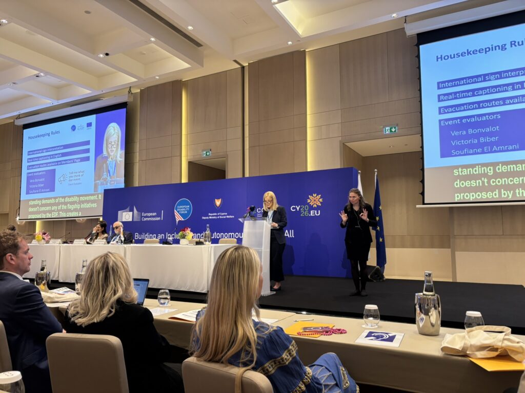 Eine Frau spricht bei der Generalversammlung des European Disability Forum in Zypern an einem Rednerpult. Daneben steht eine Gebärdensprachdolmetscherin auf der Bühne. Im Hintergrund befinden sich eine große blaue Veranstaltungswand mit Logos der Europäischen Kommission, des European Disability Forum und der zyprischen EU-Ratspräsidentschaft sowie zwei große Leinwände mit Präsentationsfolien und Live-Untertiteln. Teilnehmer*innen sitzen im Vordergrund an Tischen im Konferenzsaal.
