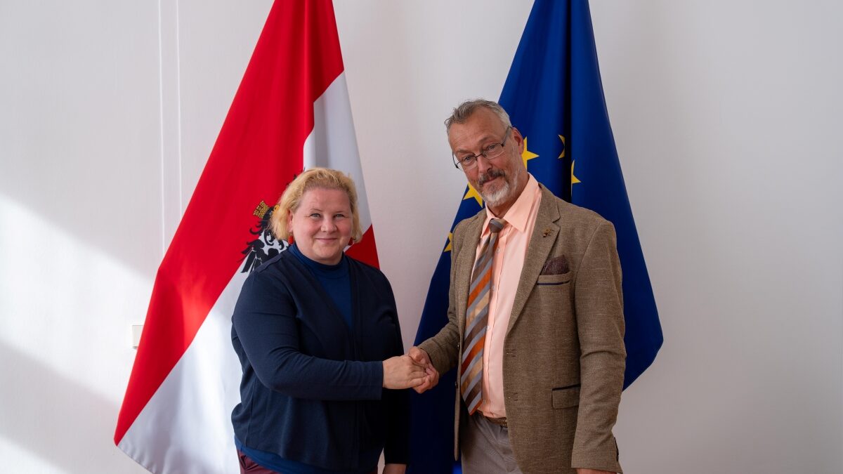Klaus Widl und Sozialministerin Korinna Schumann stehen vor einer österreichischen und einer EU-Flagge und geben sich die Hand.