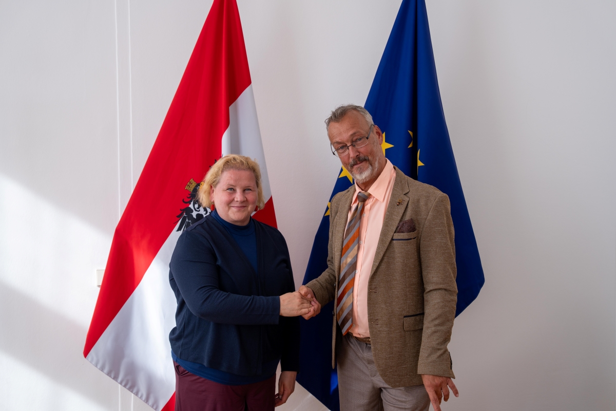 Klaus Widl und Sozialministerin Korinna Schumann stehen vor einer österreichischen und einer EU-Flagge und geben sich die Hand.