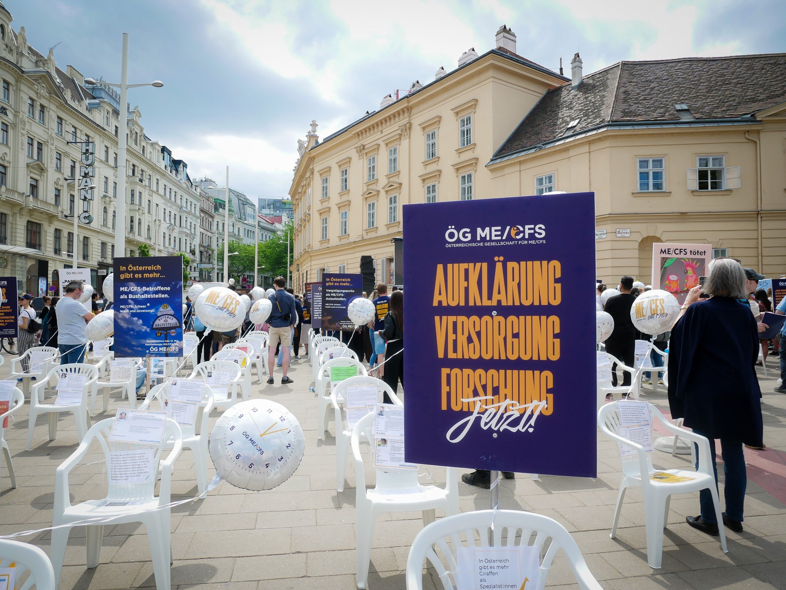 Foto der Aktion am Heldenplatz. Es sind Sessel aufgestellt, an denen Botschaften und Luftballons befestigt sind. Im Vordergrund ein Schild mit der Aufschrift "Aufklärung, Versorgung, Forschung. Jetzt"