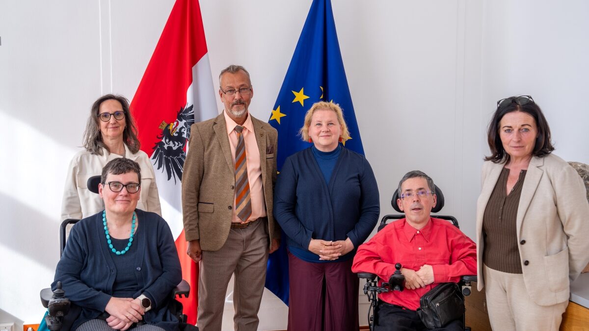 Vertreter*innen des Präsidiums des Österreichischen Behindertenrats mit Sozialministerin Korinna Schumann stehen und sitzen gemeinsam vor einer österreichischen und einer EU-Flagge.