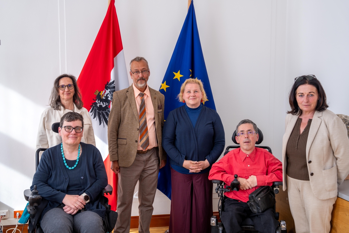Vertreter*innen des Präsidiums des Österreichischen Behindertenrats mit Sozialministerin Korinna Schumann stehen und sitzen gemeinsam vor einer österreichischen und einer EU-Flagge.