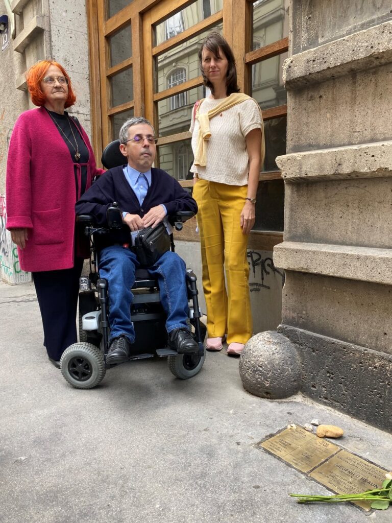 Drei Personen stehen bei der Enthüllung eines Steins der Erinnerung in der Wiener Josefstadt vor einem Hauseingang. In der Mitte sitzt Behindertenrats-Vizepräsident Martin Ladstätter im Elektrorollstuhl. Neben ihm stehen LAbg. Mag. Astrid Rompolt und NAbg. a.D. Dr. Irmtraut Karlsson. Im Gehsteig vor ihnen sind die Messingtafeln des Gedenksteins für Siegfried Braun und die Erste österreichische Krüppelarbeitsgemeinschaft eingelassen.