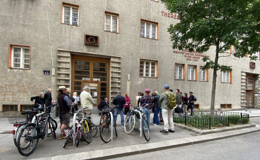 Mehrere Menschen stehen vor dem Therese-Schlesinger-Hof in der Wiener Josefstadt und nehmen an der Enthüllung eines Steins der Erinnerung für Siegfried Braun und die Erste österreichische Krüppelarbeitsgemeinschaft teil. Vor dem Gebäude sind Fahrräder abgestellt.
