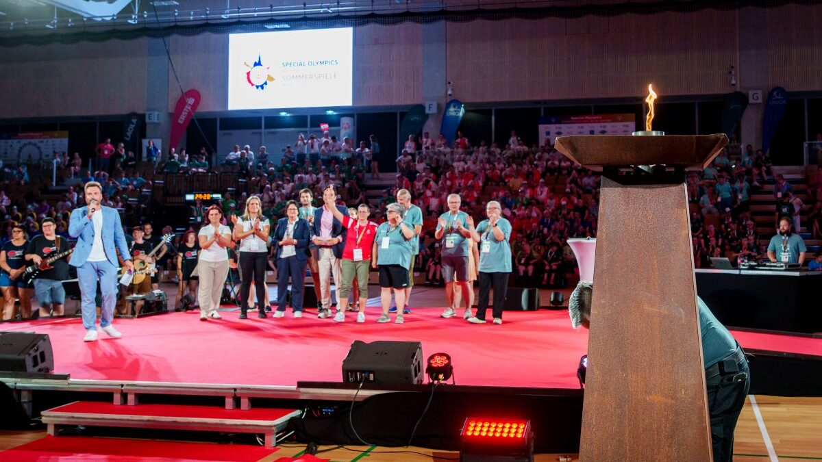 Das Special-Olympics-Feuer brennt bei der Eröffnungsfeier der 9. Nationalen Special Olympics Sommerspiele. Im Hintergrund stehen Teilnehmer*innen und Ehrengäste auf der Bühne vor einem voll besetzten Zuschauerbereich.