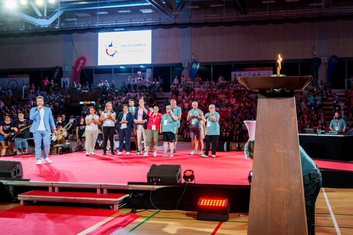 Das Special-Olympics-Feuer brennt bei der Eröffnungsfeier der 9. Nationalen Special Olympics Sommerspiele. Im Hintergrund stehen Teilnehmer*innen und Ehrengäste auf der Bühne vor einem voll besetzten Zuschauerbereich.