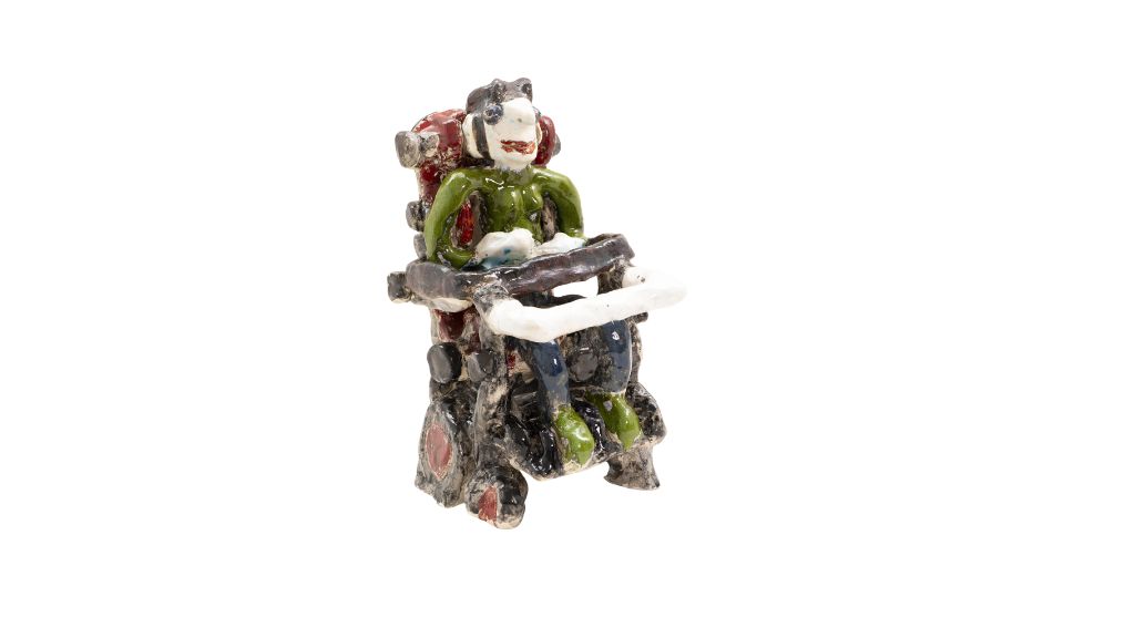 Keramikfigur „Gabi“ von Anna Magdalena Weiss (2002). Die bunt glasierte Figur zeigt eine Person mit dunklen Haaren in einem elektrischen Rollstuhl mit roter Rückenlehne. Die Person trägt ein grünes Oberteil, blaue Hosen und grüne Schuhe. Die Keramik ist bewusst grob modelliert und handgearbeitet, mit sichtbaren Strukturen und unregelmäßigen Formen.