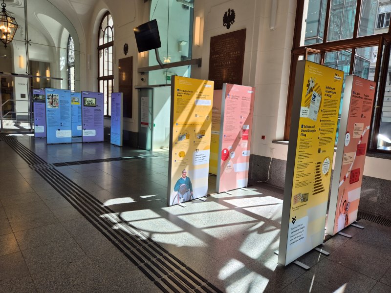 Mehrere Ausstellungstafeln zu Gleichstellung und Behindertenpolitik stehen in der Eingangshalle des Sozialministeriums in Wien. Sonnenlicht fällt durch die großen Fenster.