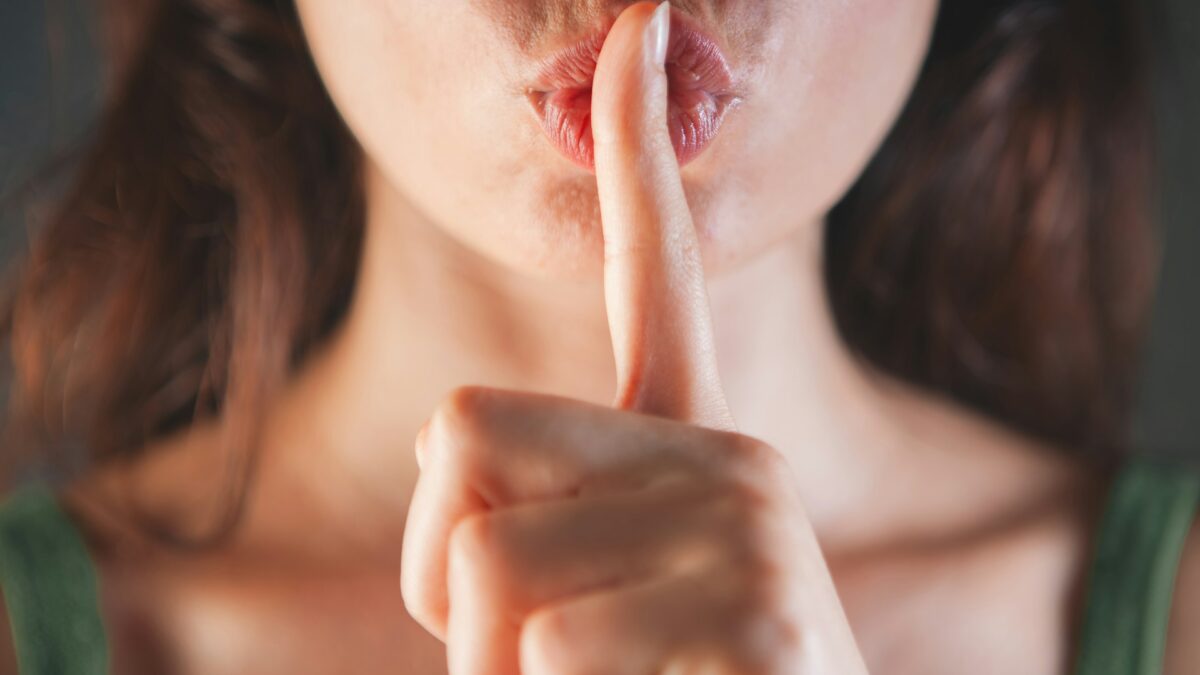 Das Bild zeigt den Mund- und Nasenbereich einer Person, die einen Finger vor die Lippen hält und damit eine Geste für Ruhe oder Stille macht. Das Gesicht ist nur teilweise sichtbar, der Hintergrund ist unscharf.