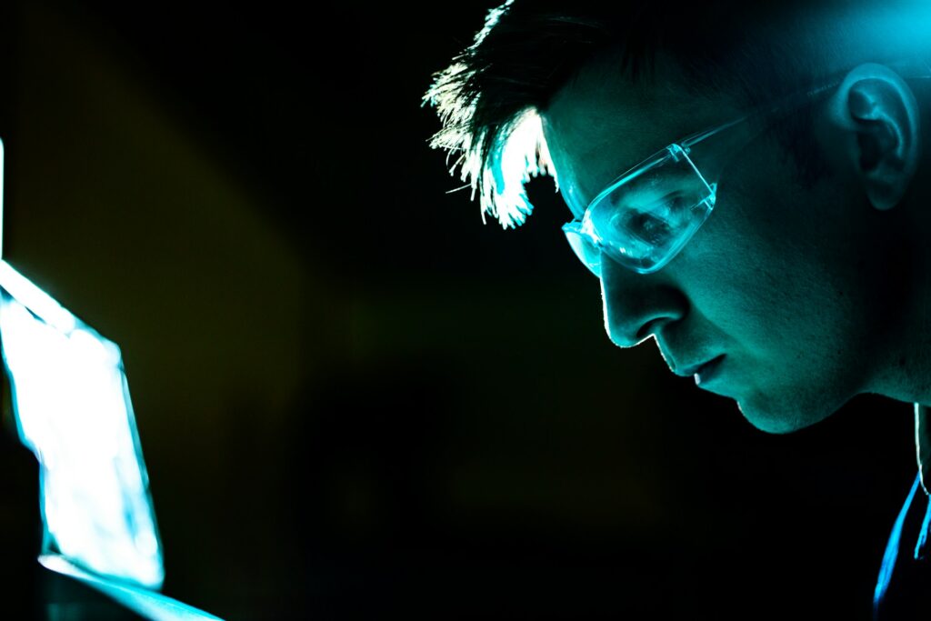 Person mit Schutzbrille blickt konzentriert auf einen hell leuchtenden Bildschirm. Das Gesicht wird von blauem Licht beleuchtet, der Hintergrund bleibt dunkel. Die Aufnahme vermittelt eine Arbeits- oder Forschungssituation im technischen Umfeld.
